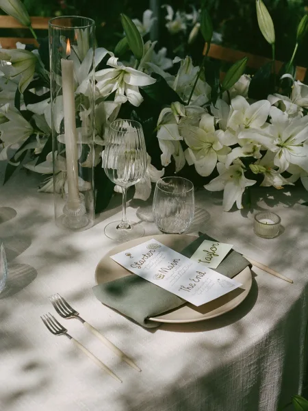 Reception tablescape with wedding place setting, white lily centerpiece, taper candles in glass vases, and place cards amid green foliage