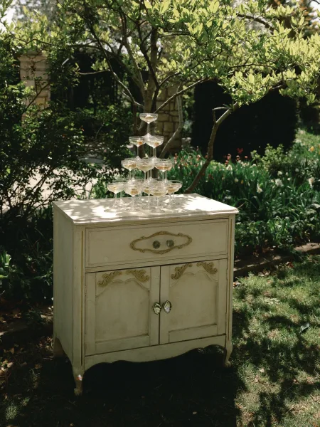 Champagne tower of coupe glasses filled with champagne atop a vintage cabinet, set in a sunlit garden with trees and a stone archway