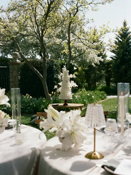 Wedding cake display on a round outdoor wedding cake table with tiered white cake and butterfly décor, lilies and candles on a garden lawn