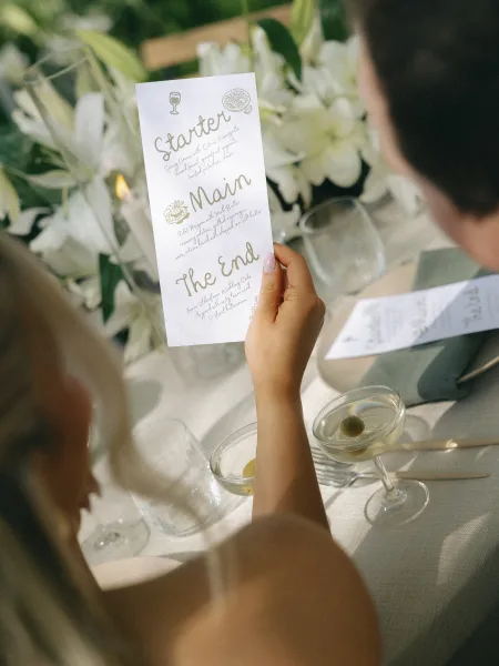 Wedding menu card with wedding menu design in calligraphy lettering beside white floral centerpiece, candle, glassware, and gold flatware on reception table