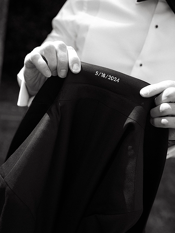 Wedding suit detail showing a black tuxedo jacket with embroidered wedding date, white dress shirt, and cufflinks against a dark interior background