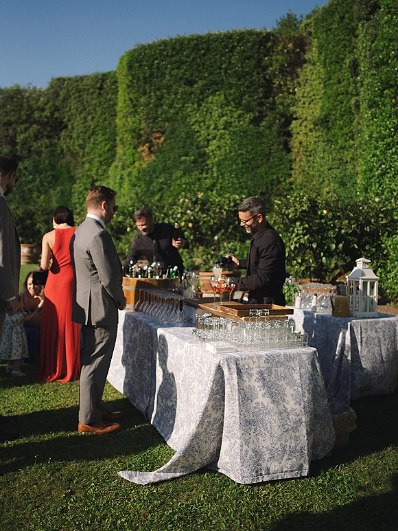 Wedding bar setup with champagne coupe glasses and liquor bottles on patterned tables, rustic barrel and white lanterns on a garden lawn