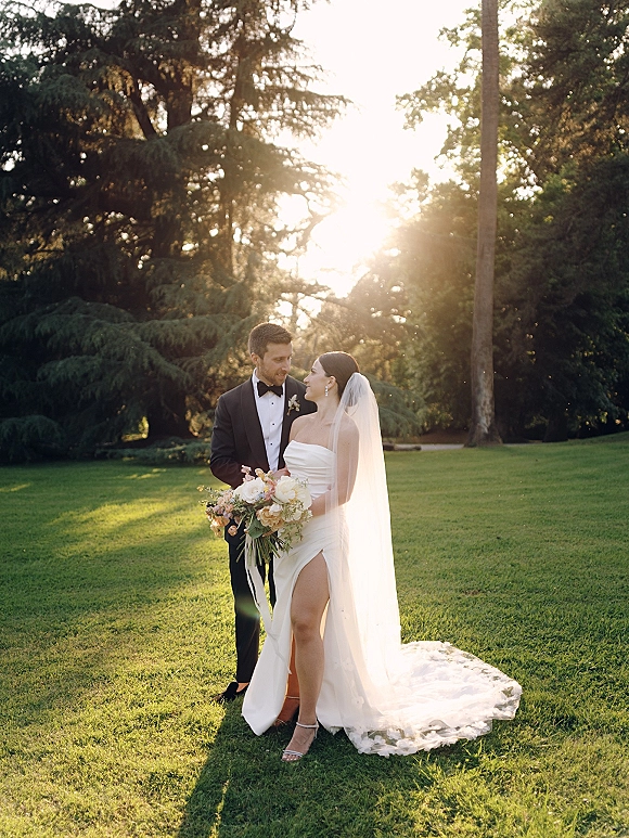 Couple portrait of bride holding a rose bouquet beside groom in black tuxedo, long veil and slit gown, backlit on a sunny garden lawn