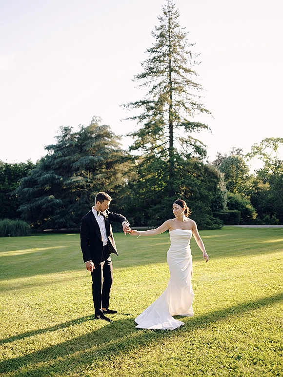Couple portrait of bride and groom holding hands, her strapless dress flowing as he wears a tuxedo on a garden lawn with tall trees.