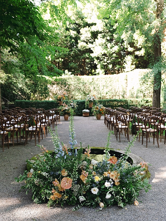Ceremony aisle decor with wooden chairs lining a gravel path, leading to a fountain framed by ground florals and garden trees in dappled sunlight