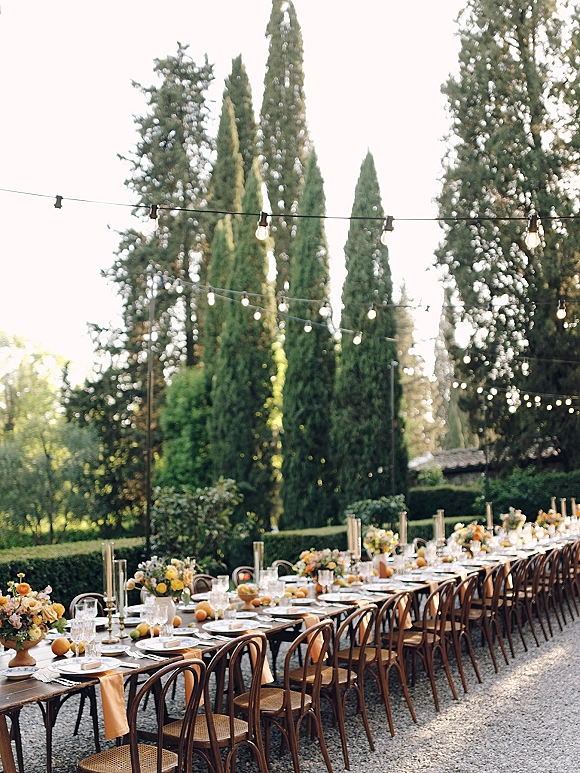 Outdoor reception tablescape with long banquet tables under string lights, yellow florals, taper candles, citrus accents, and evergreen garden backdrop