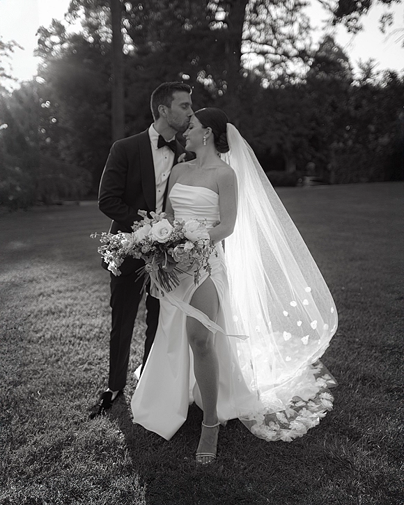 Couple portrait of groom kissing bride’s forehead as she holds a bouquet, her long veil and strapless dress glowing on a sunny lawn