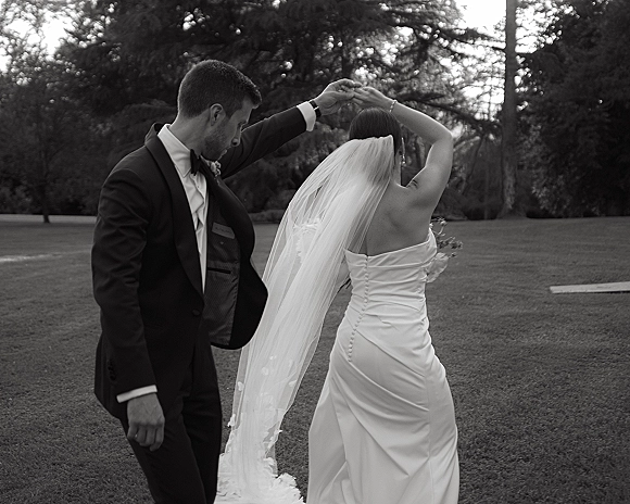 Wedding couple dancing as the bride and groom twirl, her long veil trailing behind in a garden park with trees in the background