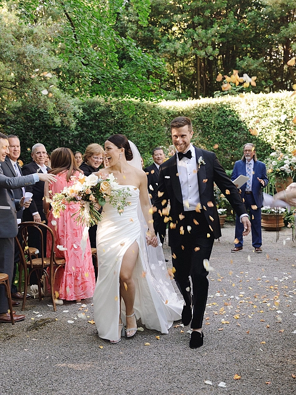 Wedding recessional of newlyweds walking down aisle, holding hands as guests toss flower petals along a garden hedge-lined path