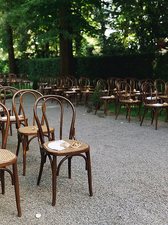 Ceremony seating with bentwood chair rows along a gravel aisle, each set with a paper fan, program, and floral boutonniere in a garden setting