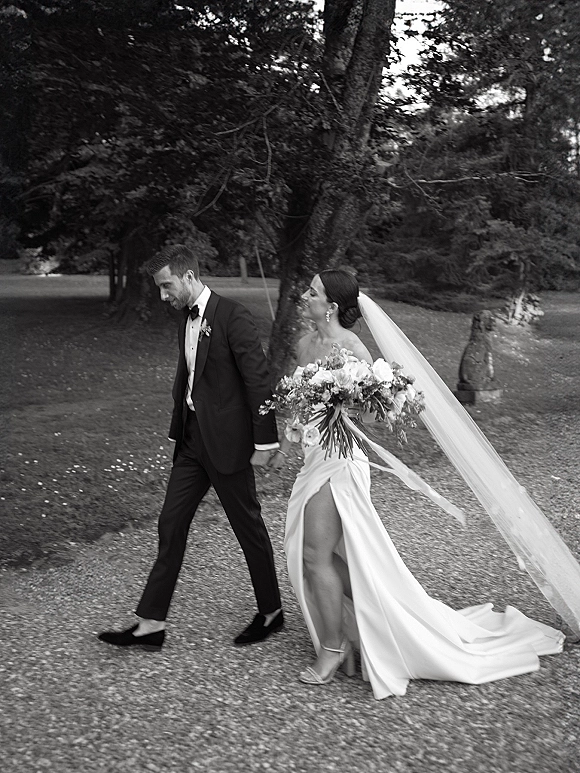 Couple portrait of bride and groom walking hand in hand, bouquet and long veil trailing, on a tree-lined garden gravel path