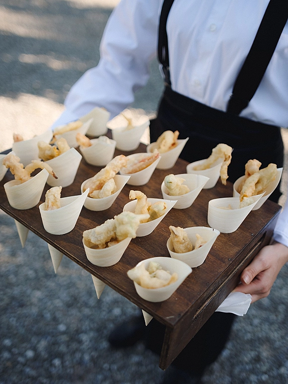 Wedding passed appetizers on a wood serving tray in small cups, held by a server in suspenders on outdoor pavement during cocktail hour