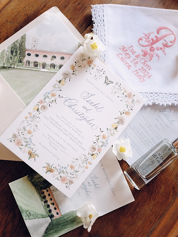 Invitation suite with floral wedding invitation, RSVP card, envelope liner and lace monogram handkerchief beside perfume on a wood tabletop