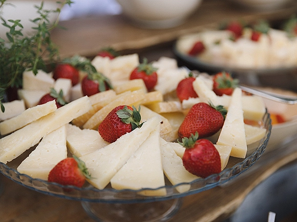 Wedding dessert table featuring a cheese and strawberry platter on a glass pedestal stand with herb garnish, bowls, and serving tongs on a wood buffet table