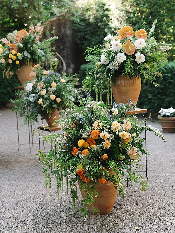Ceremony floral arrangements in large terracotta urns with orange anthurium, roses, ranunculus, and trailing greenery along a garden path