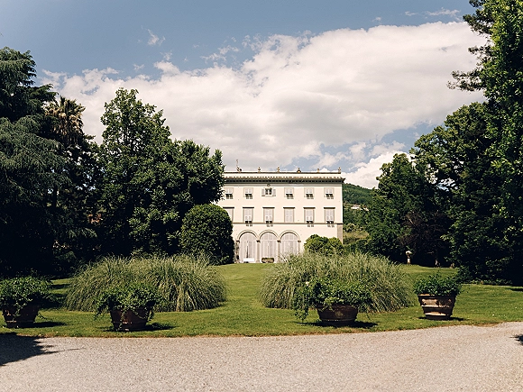 Wedding venue exterior with a white villa facade, potted grasses accenting the gravel drive, set against lawns, trees, and hills under blue sky