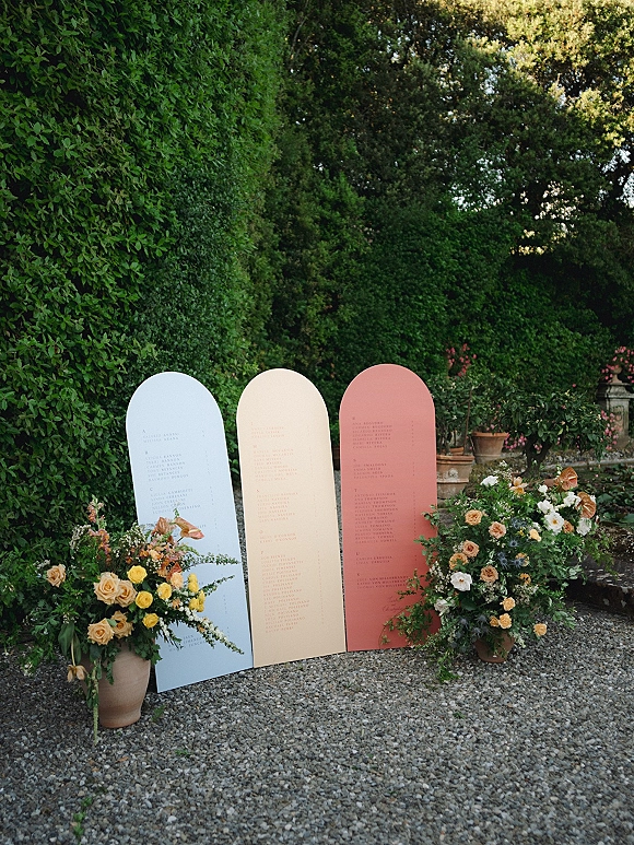Wedding seating chart with three arched panels and rose florals in terracotta pots along a gravel garden path with hedges