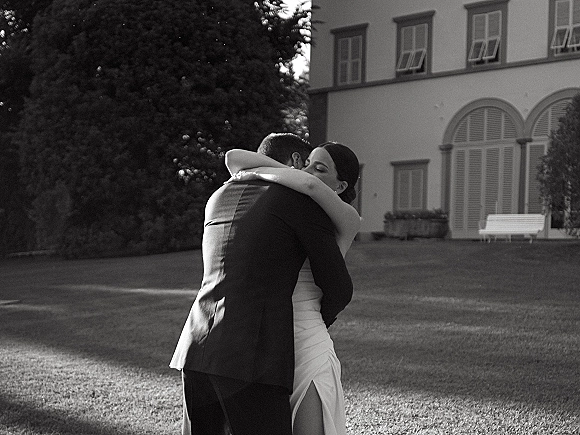 Couple embrace in a wedding hug photo, bride in a strapless gown with earrings and groom in suit on lawn by estate windows