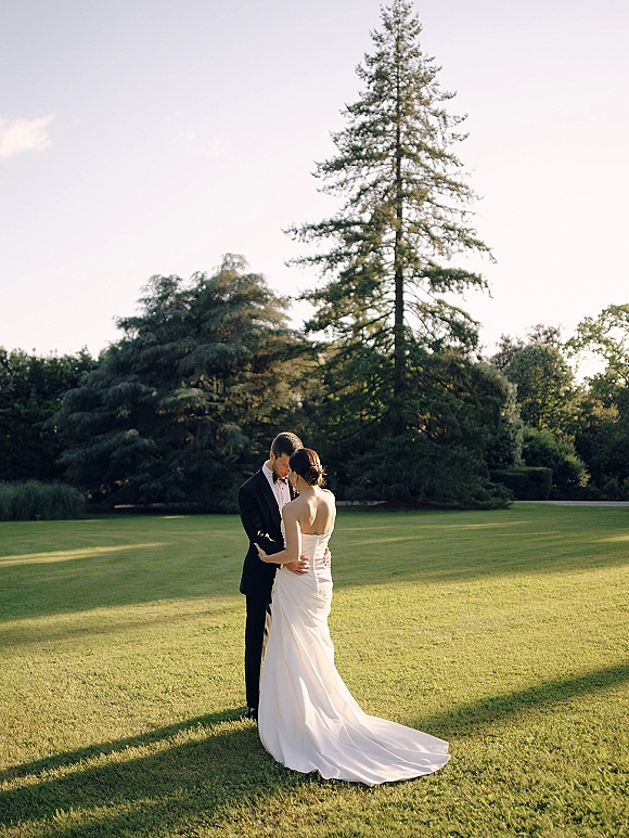 Couple portrait of bride and groom embracing with a forehead touch, her strapless dress train and his tuxedo on a grassy lawn by pines