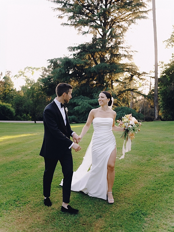 Couple portrait of bride and groom walking hand in hand on a green lawn, bride in strapless slit dress holding bouquet with ribbon and veil