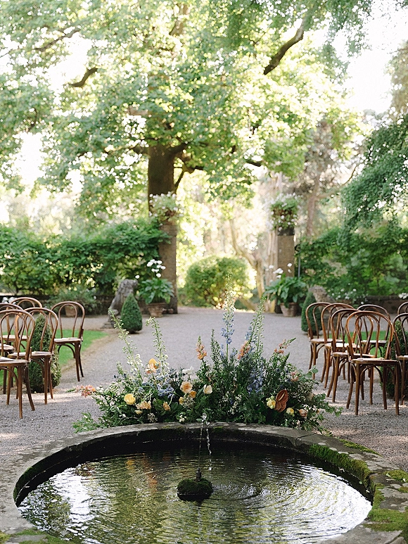 Outdoor ceremony setup with wood chairs lining a gravel aisle and a low floral ground arrangement beside a stone garden fountain
