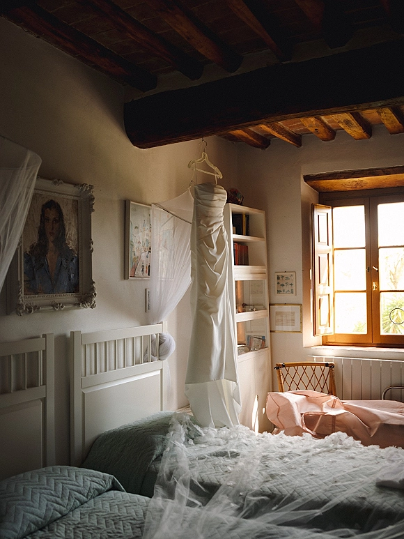 Wedding dress hanging on a hanger, strapless satin gown by a sunlit bedroom window, with tulle veil draped across the bed