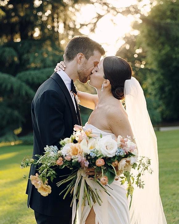Wedding kiss portrait of bride and groom kiss, her veil draped over shoulder and bouquet held close in a sunlit garden setting