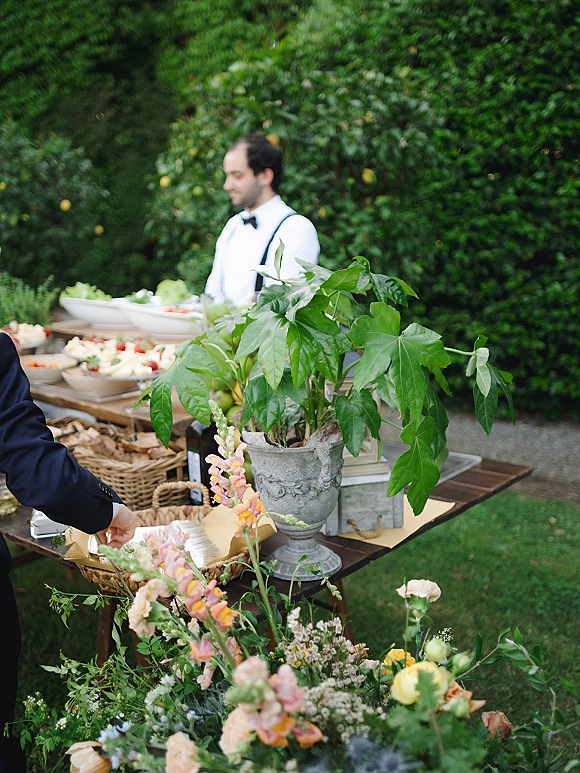 Wedding catering table set as an outdoor wedding buffet with food platters, wicker baskets, and a floral accent amid garden hedges