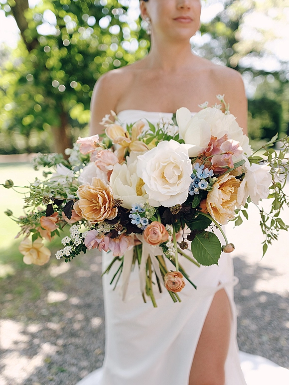 Bridal bouquet with white roses and peach blooms, touched with blue accents and greenery, held by bride in strapless gown on a garden path