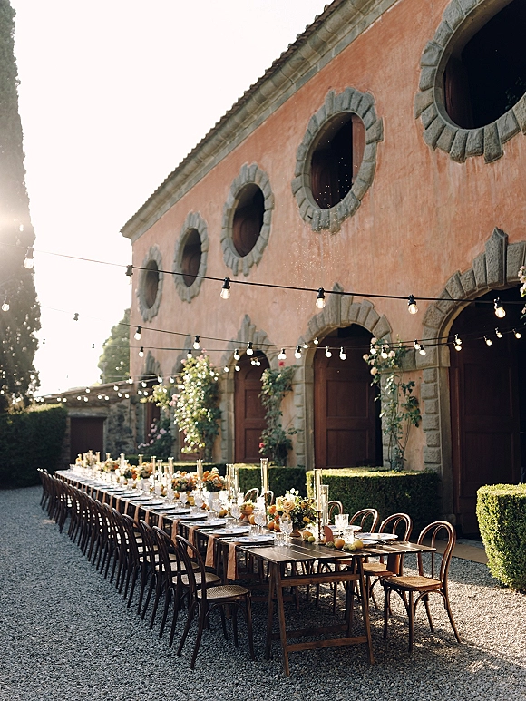 Outdoor reception tablescape with a long banquet table wedding setup, floral centerpieces, taper candles and string lights in a gravel courtyard