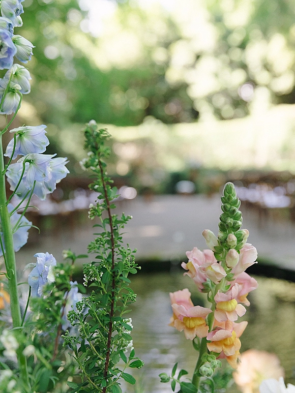 Wedding ceremony aisle lined with delphinium and snapdragons, soft pastel ground florals leading toward a wooden bridge by a pond