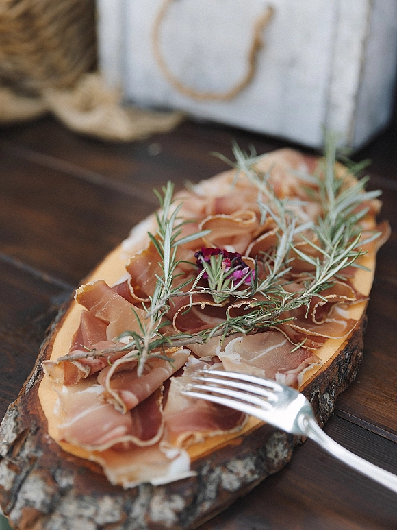 Wedding appetizers on a charcuterie wedding appetizer board with prosciutto, rosemary sprigs, fork, and dried flowers on a wood table