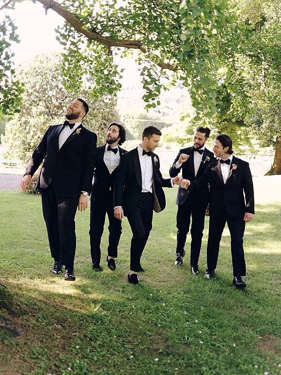 Groomsmen portrait of the wedding party men walking and laughing in black tuxedos with bow ties on a sunlit garden lawn