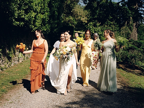 Bridal party walking along a sunlit garden path, bride with bridesmaids carrying bouquets in mismatched dresses amid trees and stone wall