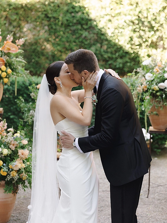 Wedding kiss as bride and groom embrace, her long veil flowing beside terracotta pots and floral pillars on a garden stone patio
