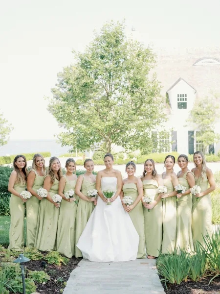 Bridesmaids group photo of a bride with bridesmaids in sage green dresses holding white bouquets on a garden lawn by a white house exterior