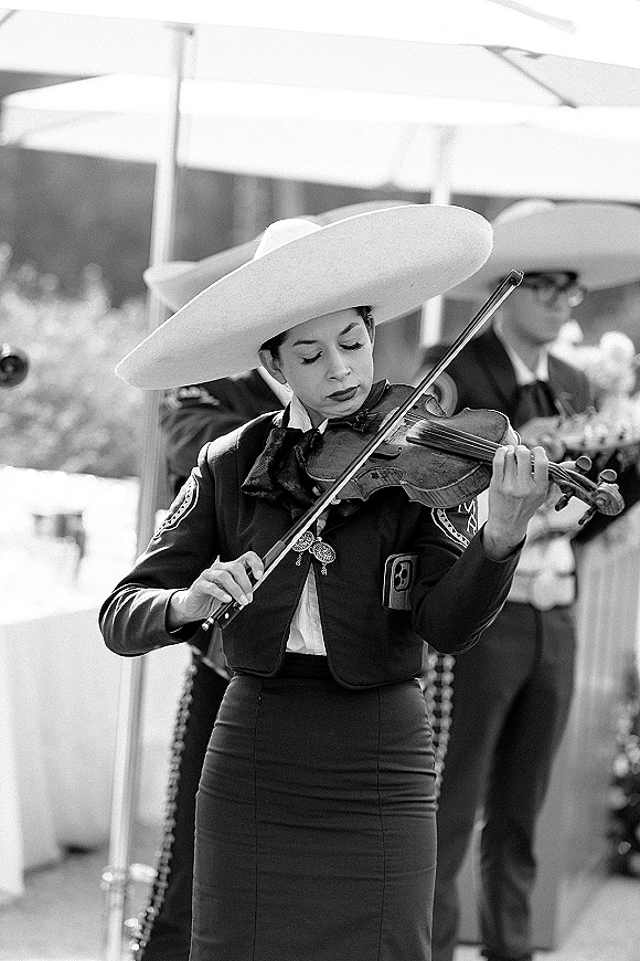 Wedding mariachi band mariachi violinist in a charro suit playing violin with bow on a patio, umbrellas behind and musicians blurred