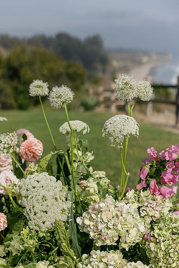 Wedding floral arrangement with ceremony aisle flowers featuring white allium and hydrangea on a lawn beside coastal cliffs and ocean view