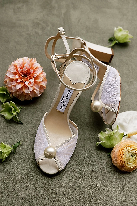 Bridal shoes in a wedding shoes flat lay, white ankle-strap sandals with pearl embellishments, flowers, and box on stone surface