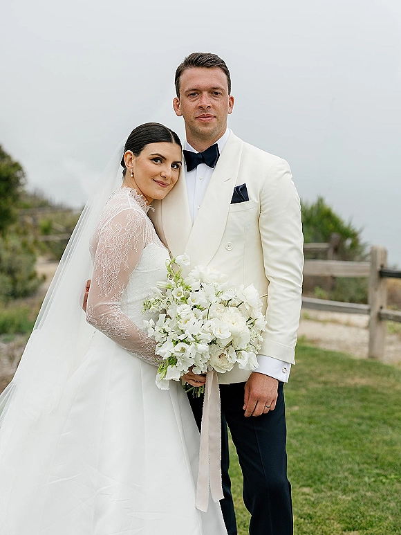Couple portrait of bride leaning on groom, holding a white bouquet, with veil and lace sleeves on a lawn by a wooden fence under gray sky