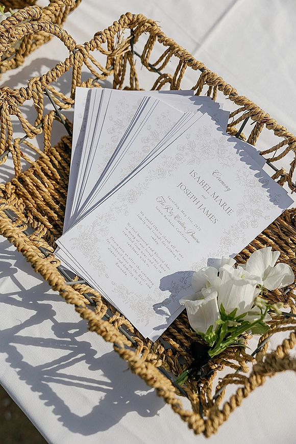Wedding invitation suite with wedding ceremony program, floral illustration border and white flowers in a woven rattan basket on linen in sunlight shadows