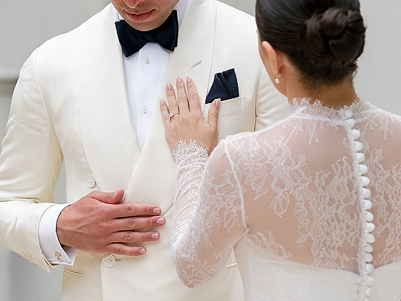 Wedding couple portrait with bride hand on groom chest, engagement ring visible beside lace sleeve dress and white tuxedo jacket on neutral wall
