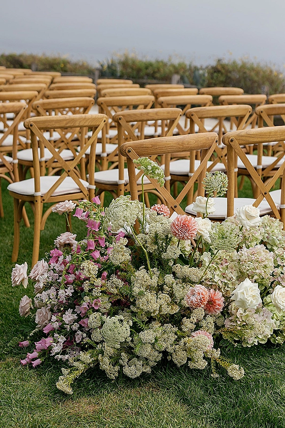 Ceremony aisle decor with wedding ceremony chair rows of wood crossback chairs and ground florals on a coastal lawn with ocean backdrop