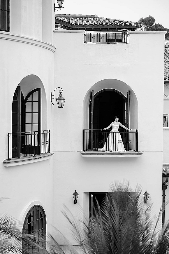 Bridal portrait of a bride on balcony in a minimalist bridal gown, looking away beside wrought iron railing at a stucco villa façade