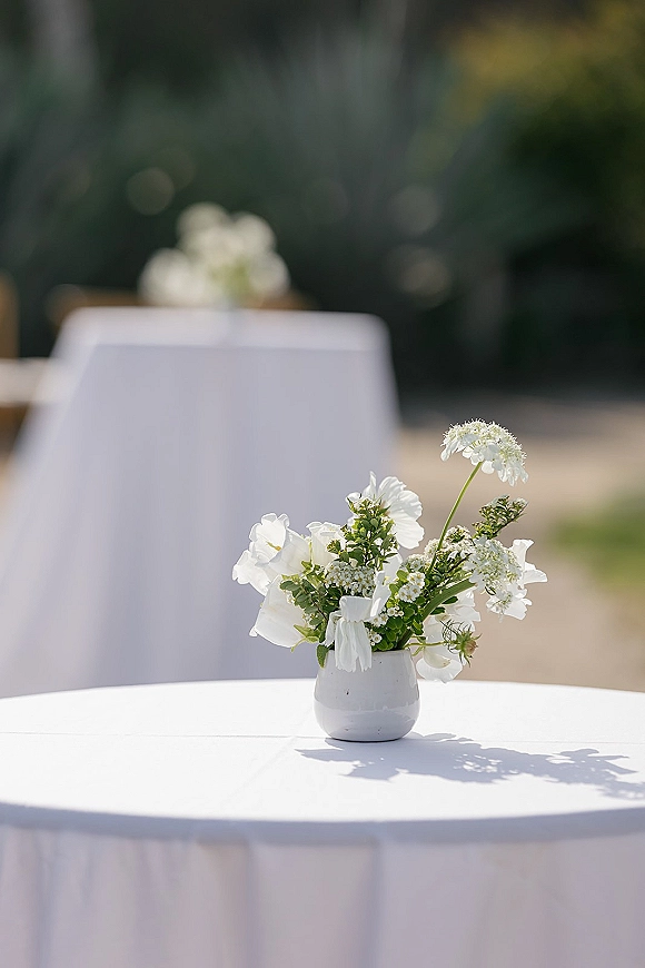 Wedding table centerpiece with white flowers in a bud vase tied with a white ribbon on a round table, outdoor greenery behind
