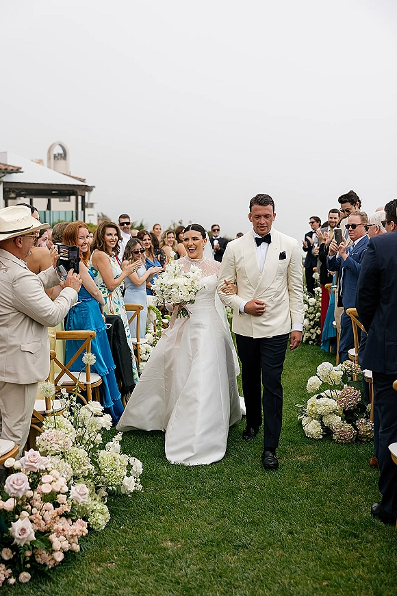 Wedding recessional as bride and groom walk the aisle, bouquet and veil flowing, guests cheering beside floral arrangements on a lawn