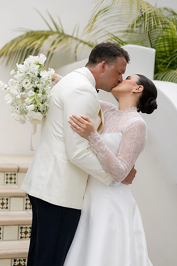 Wedding kiss as bride holds a white bouquet, lace long sleeves and pearl studs, groom in white tuxedo and black bow tie by palms on stucco stairs