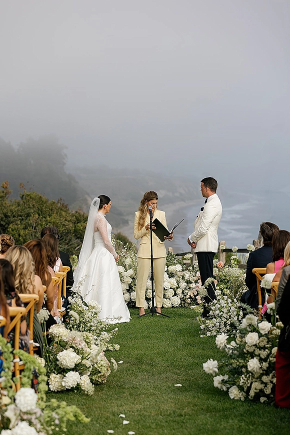 Wedding ceremony with bride in long veil and groom in tuxedo as officiant reads vows on a foggy cliffside lawn by the ocean