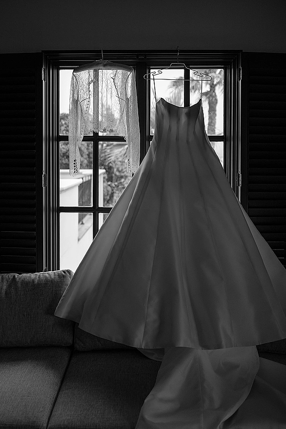 Wedding dress hanging on a hanger by window shutters, paired with a lace bridal jacket, with couch and trees softly visible beyond
