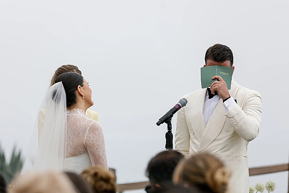 Wedding vows as groom reads vows into a microphone from a booklet while bride in a long veil listens during an outdoor ceremony with guests behind